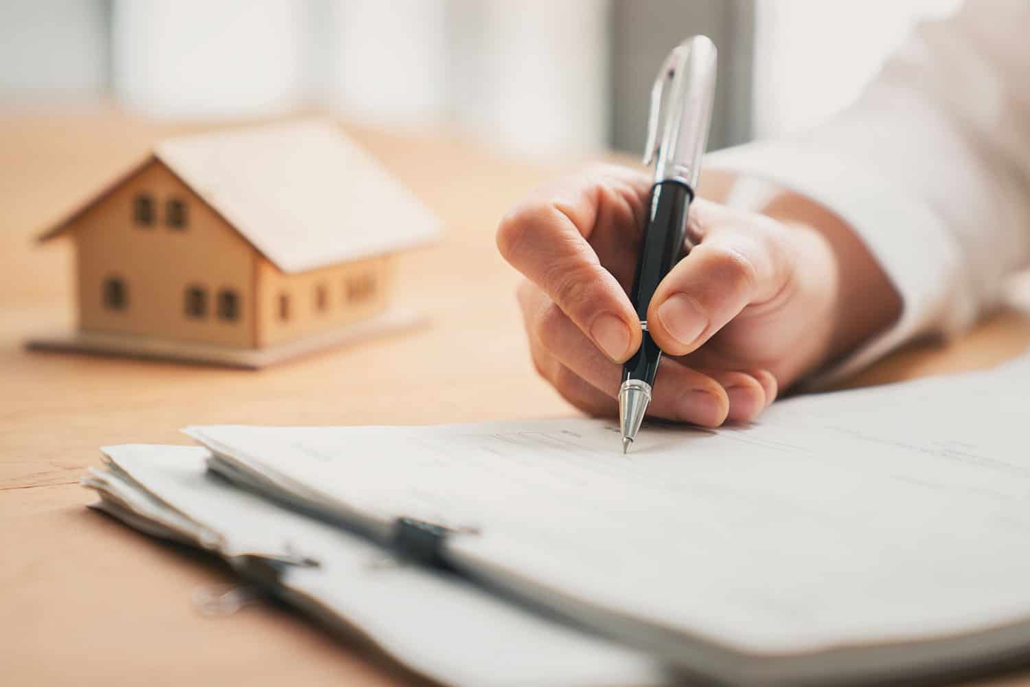photo close-up of someone’s hand, writing with a biro pen on paperwork with a model house in the background