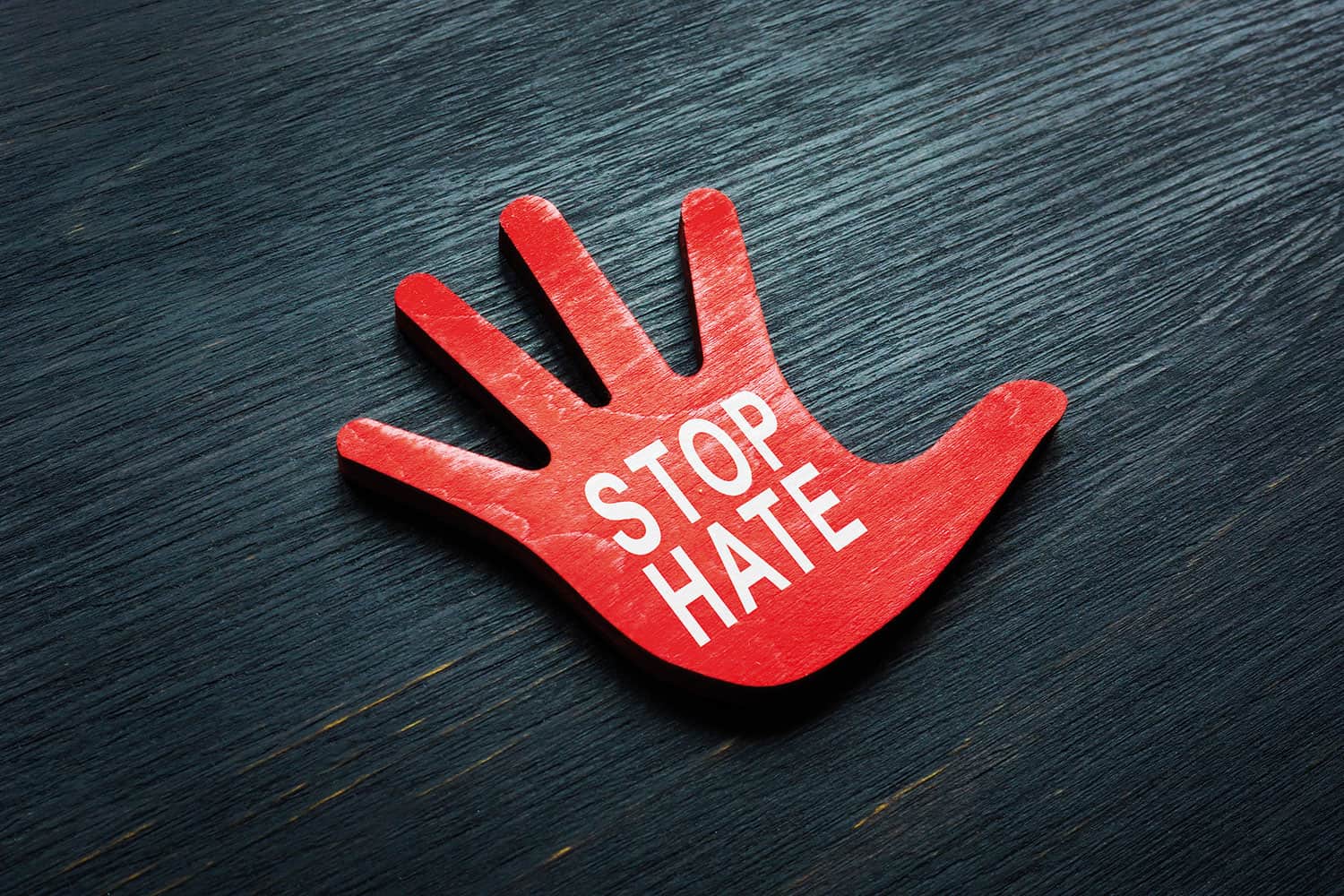 photo of 2D, flat, red-painted wooden hand with fingers spread, with words ‘STOP HATE’ in white, on dark table top background