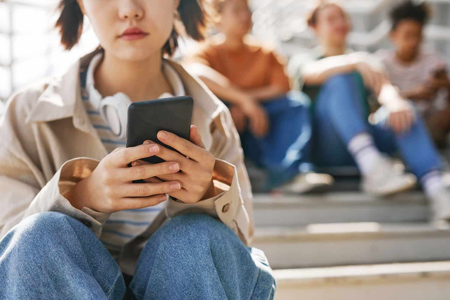 photo of young teen in casual clothes sat on a light-filled stairwell, looking at her mobile phone, which is the focus of the pic