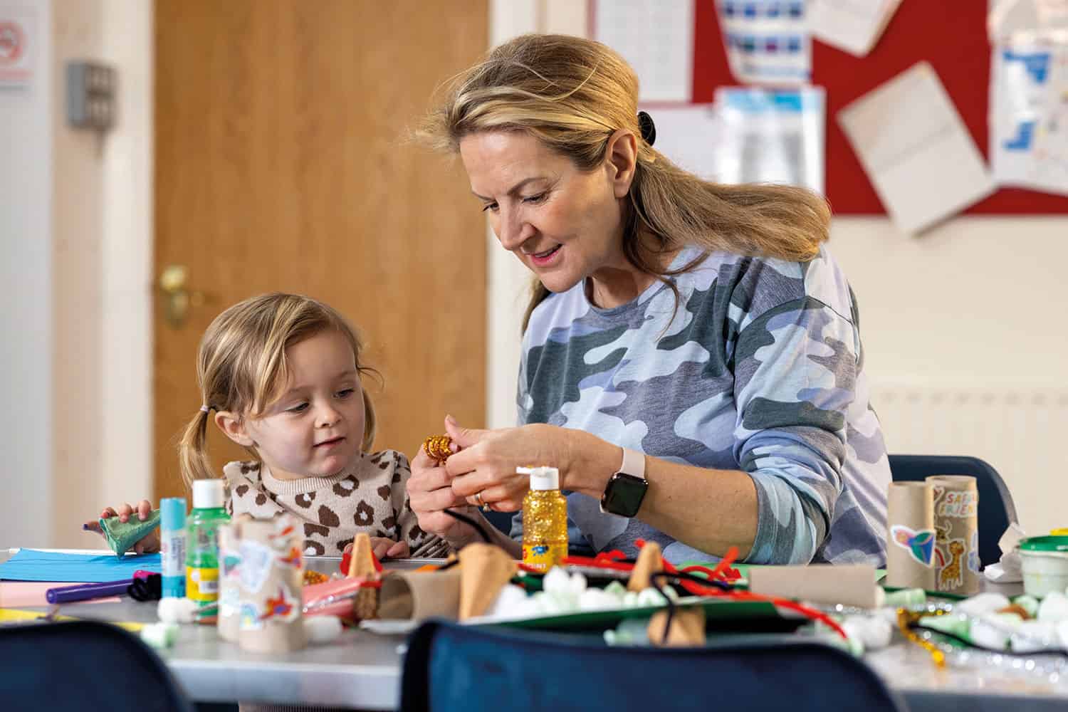 mum sitting beside pre-school daughter, playing with paper, glue and craft materials