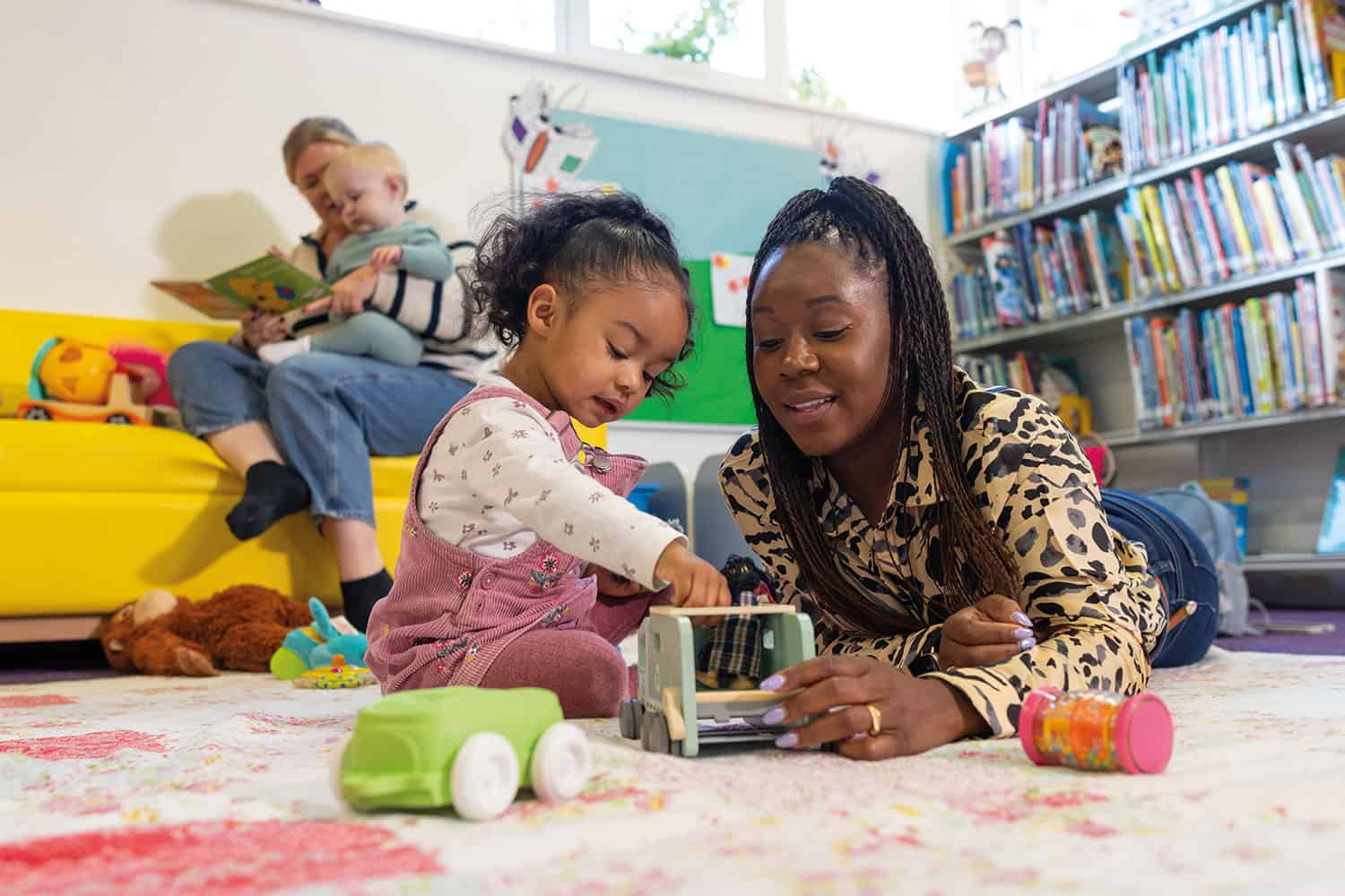 photo of mum lying on floor playing with pre-school daughter with another mum in the behind her to the left, sitting on a yellow sofa reading a book to her baby, with a book shelf and colourful display board in the background to the right.
