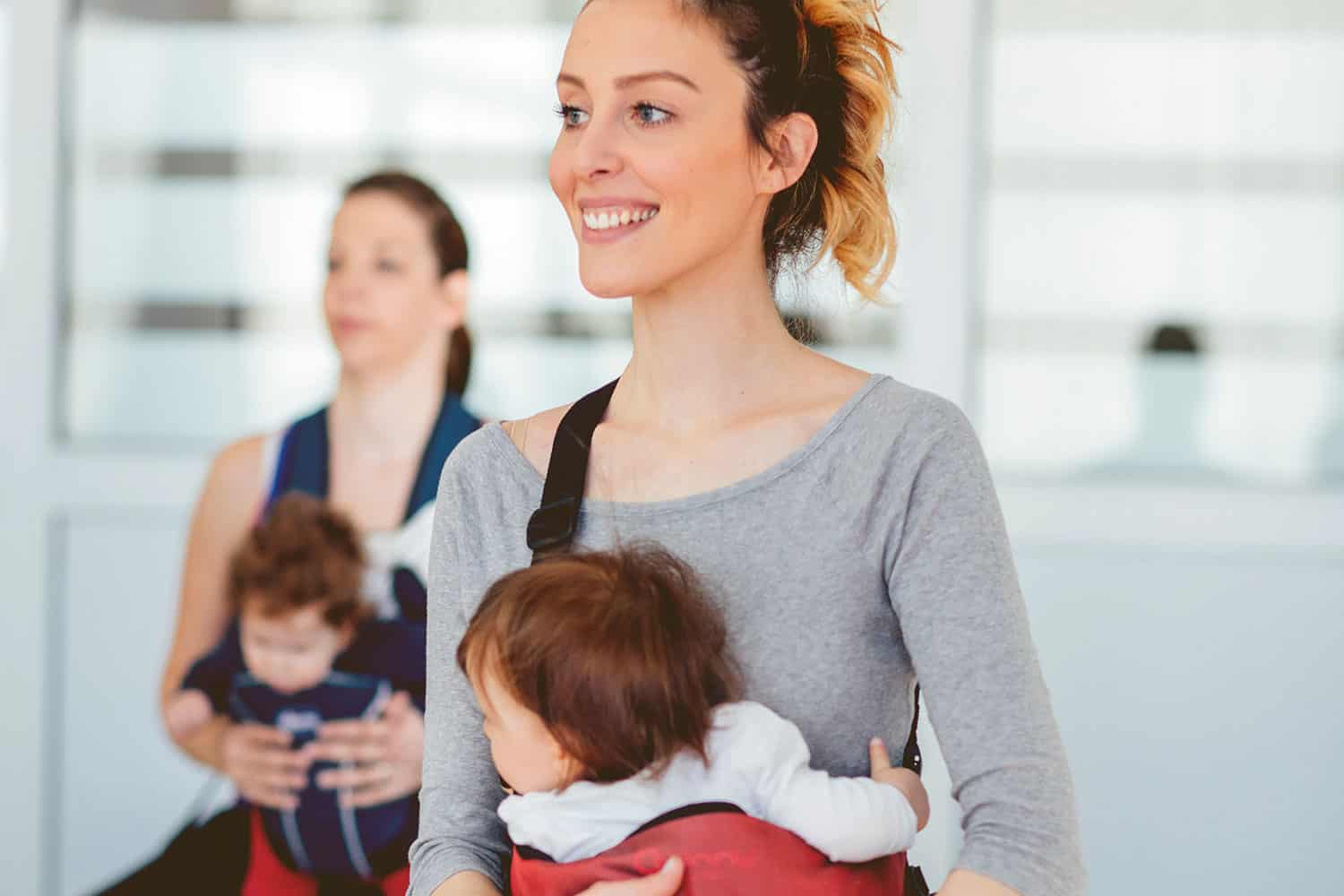 photo of smiling mum doing exercises with baby strapped to her front in a sling, and another mum and baby in the background