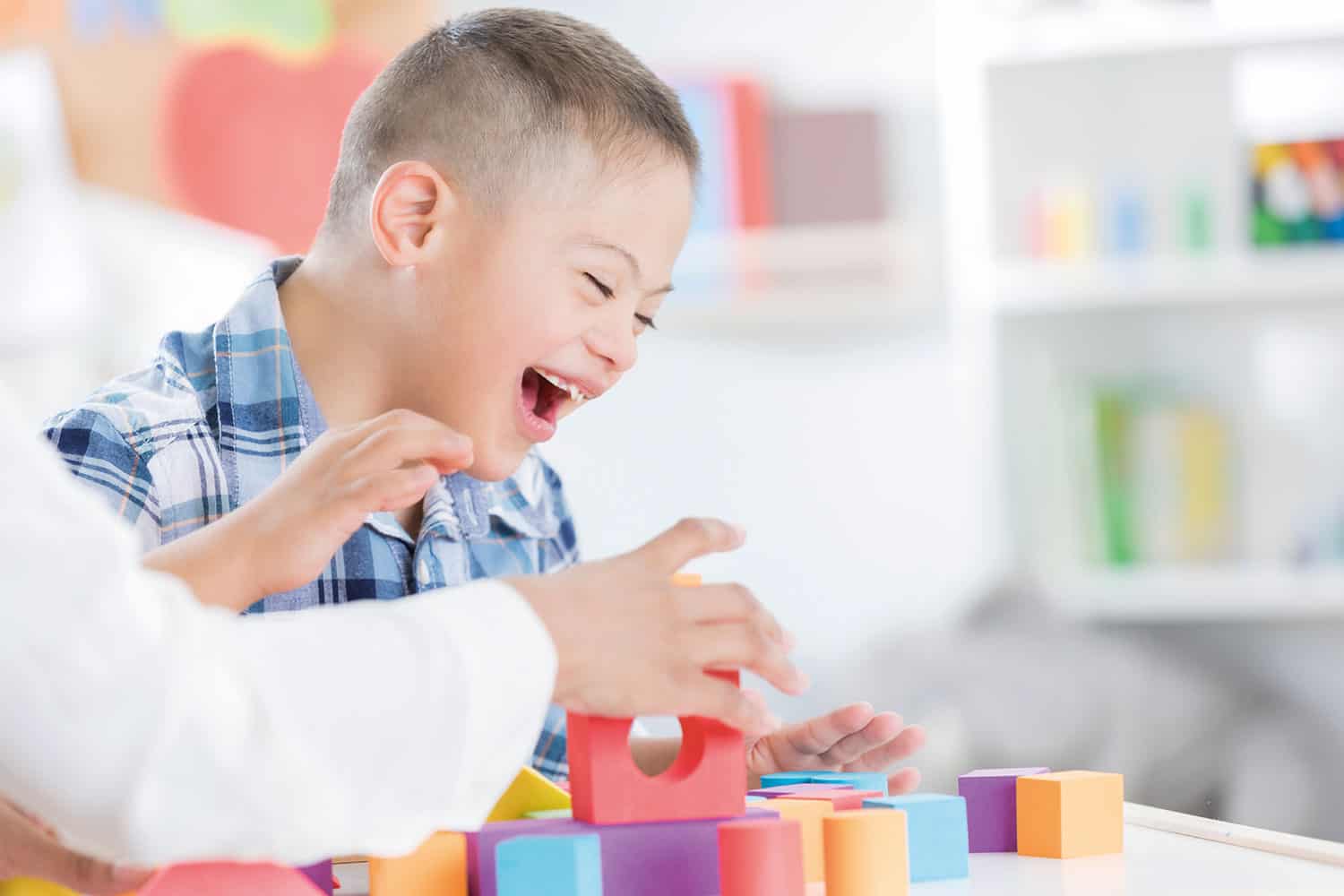 photo of young boy with Downs Syndrome laughing as he plays with shapes and building blocks on a table