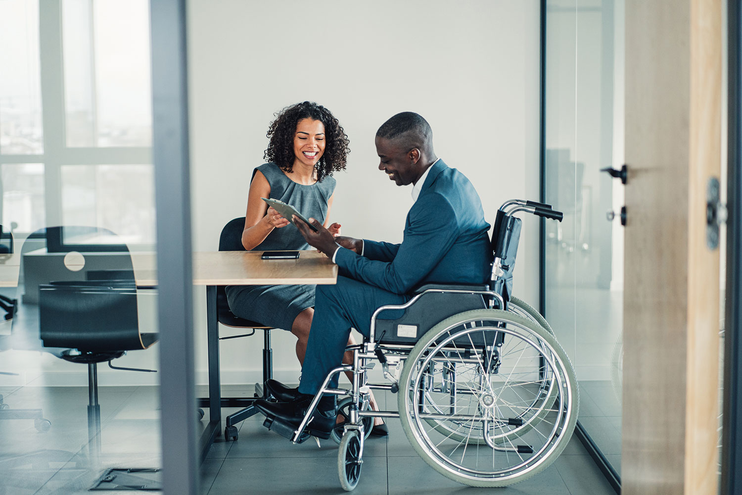 photo of a young man, a wheelchair user, sitting at an office desk with a colleague, both looking at a tablet