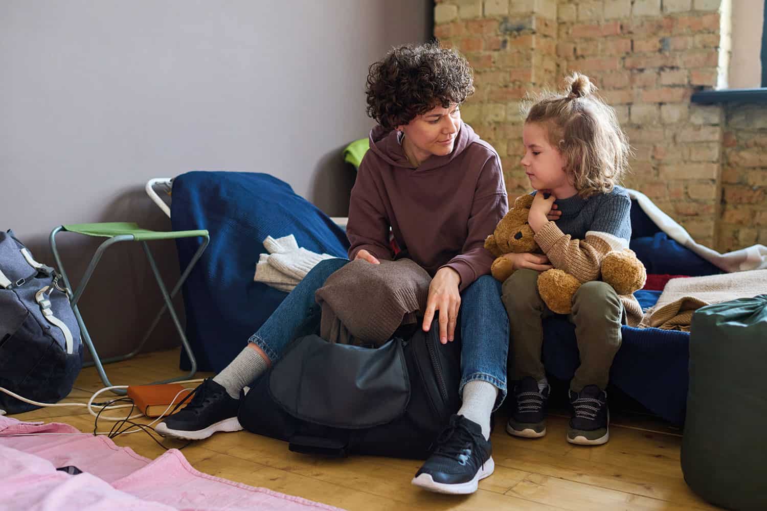 photo of mum and daughter sat on a bed, with the former holding a bag and the latter a teddy bear