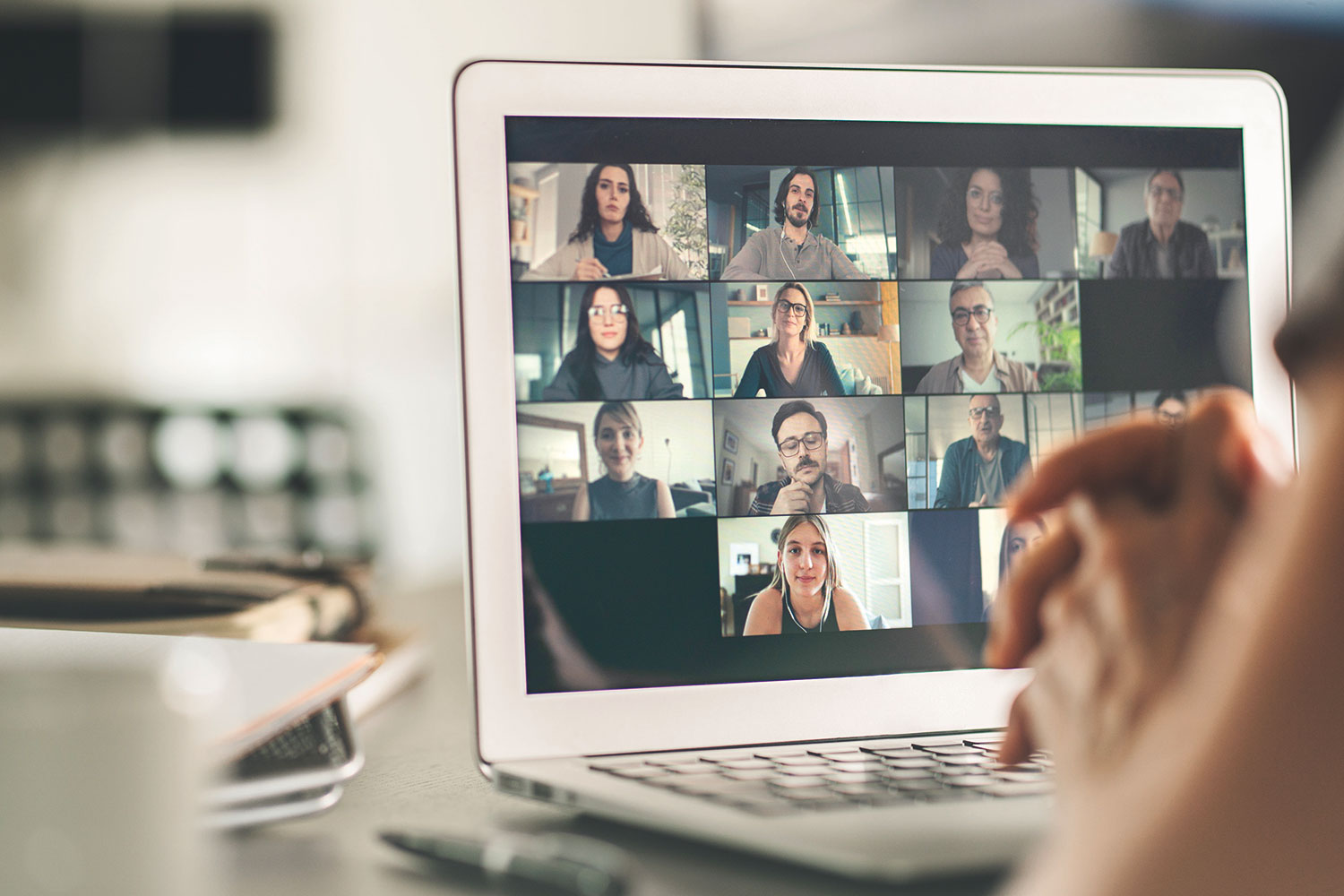 photo of a laptop screen showing people having an online meeting, taken over the shoulder of someone sitting at a desk