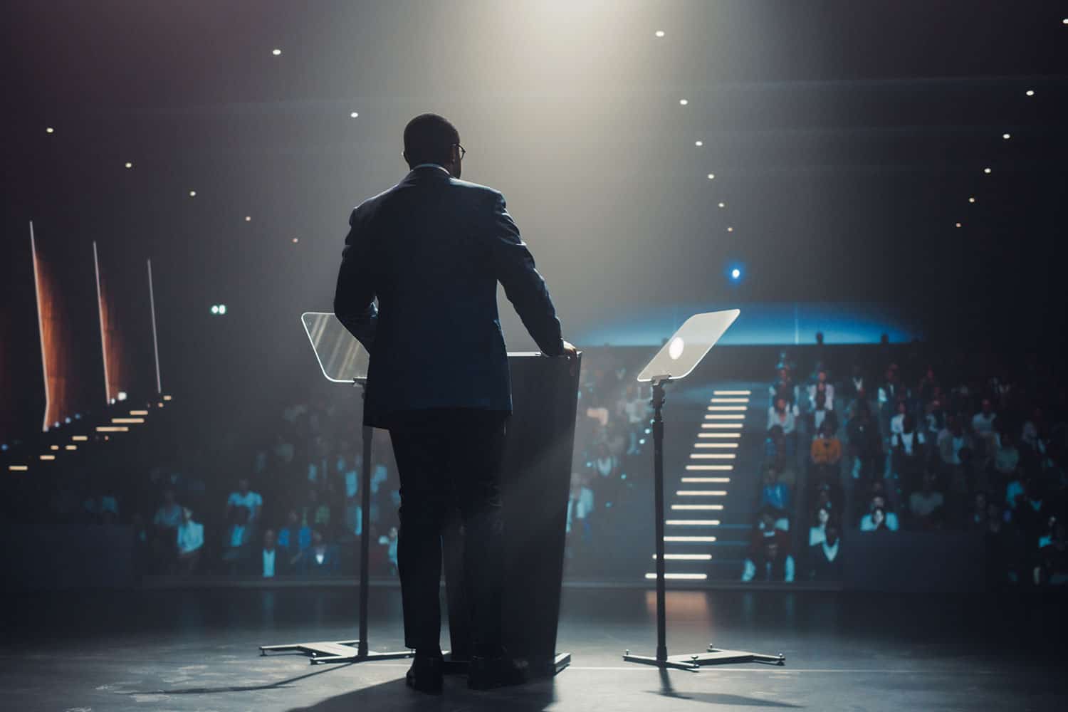 photo from behind of a speaker on a stage behind a podium with autocue screens to left and right
