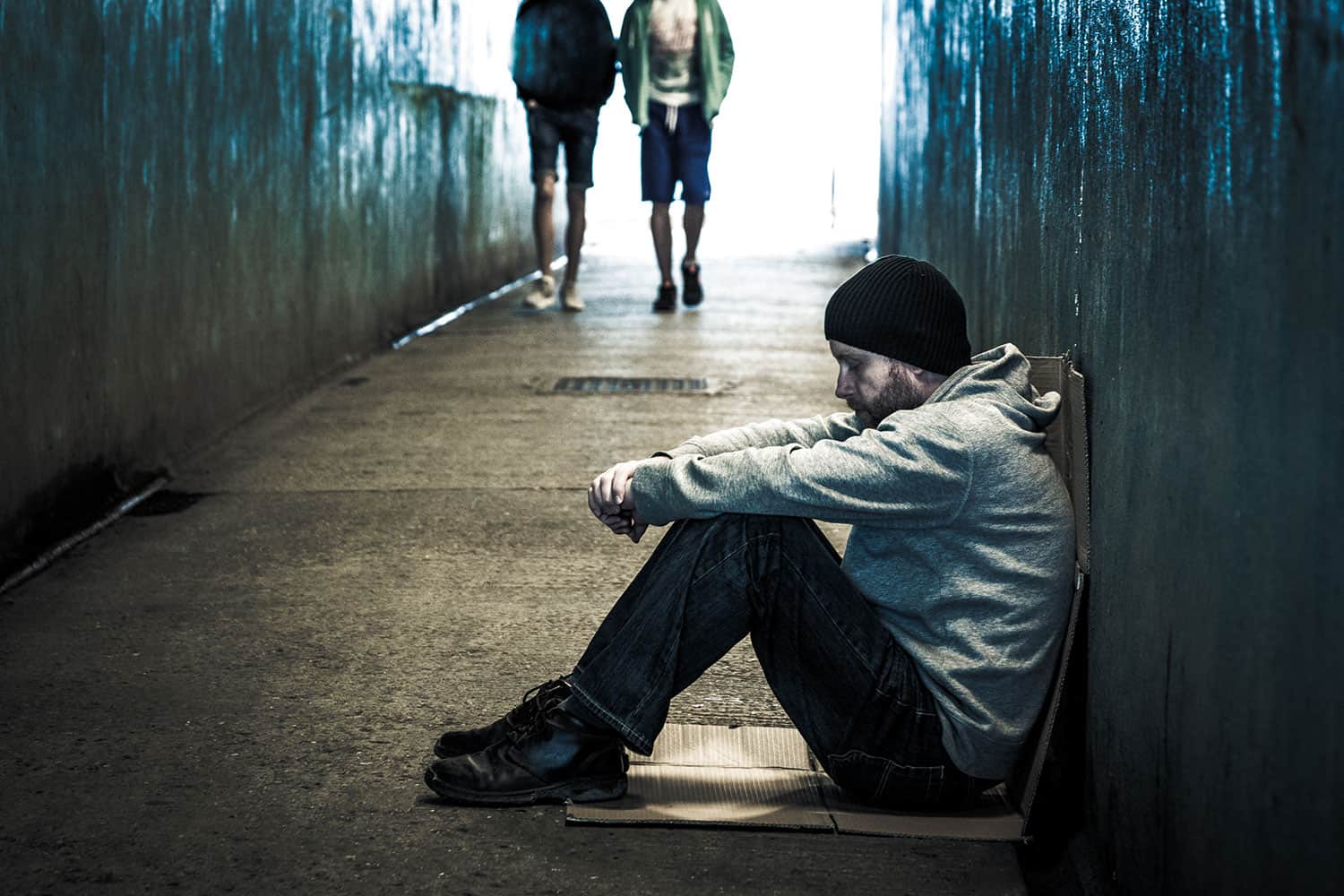 photo of homeless young man sitting on cardboard in a dark pedestrian subway with two people walking towards him from the light