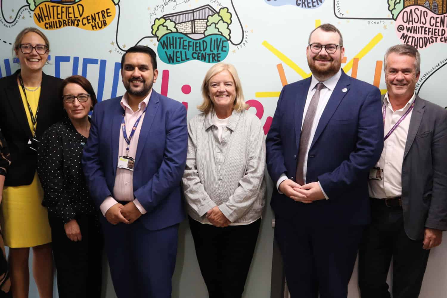 photo of Baroness Louise Casey (centre), Chair of the Independent Commission on Adult Social Care, and LGA Chair Cllr Eamonn O’Brien (right of centre) with colleagues from Bury Council, against a wall with colourful writing and graphics of local support and activity centres