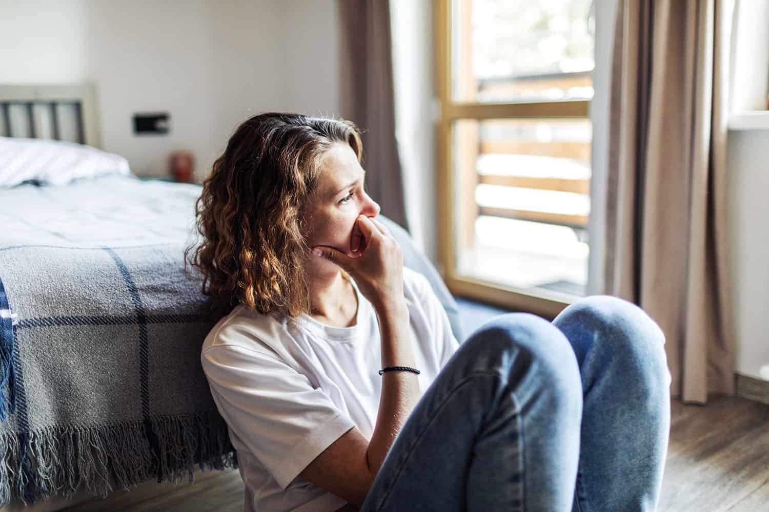 woman looking pensive, sitting on floor with her back to a bed and knees drawn up, with a window in the background