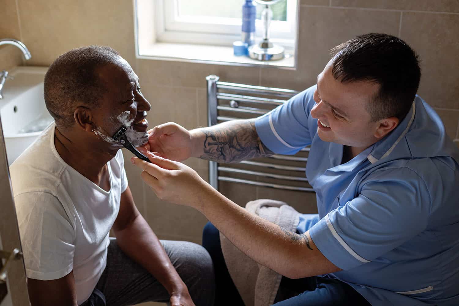 young male carer in blue uniform helping older man shave in the latter’s bathroom at home