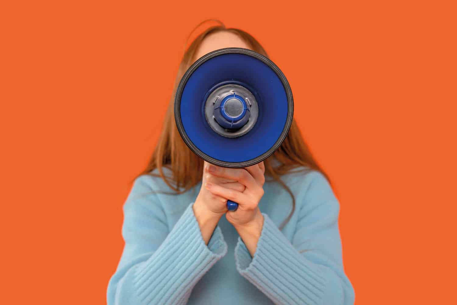 of woman with long reddish-brown hair wearing a light blue jumper holding a dark blue megaphone so it completely obscures her face, on a bright orange background